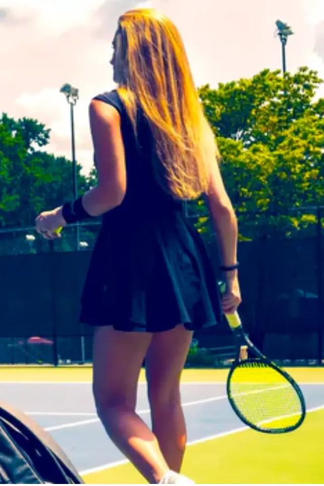 Female tennis player in a black dress holding a tennis racket, standing by the court with an ADV bag nearby