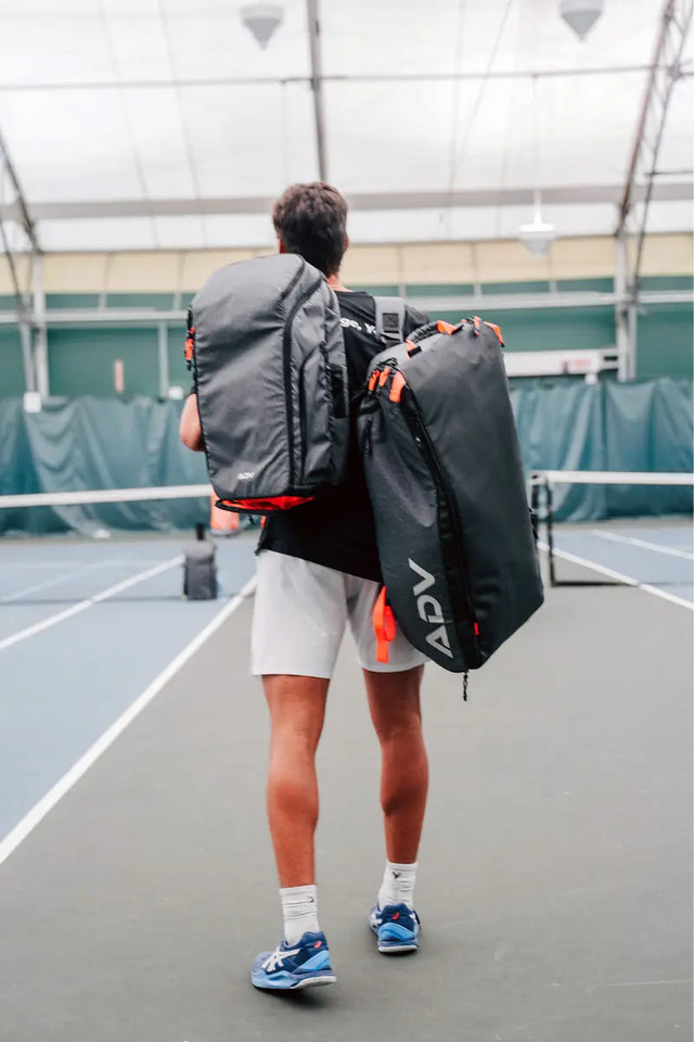 Player walks onto an indoor court carrying two black and orange ADV tennis bags.
