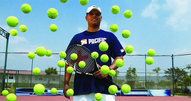 Tennis coach Braen Aneiros holds a racquet on court amid floating tennis balls.