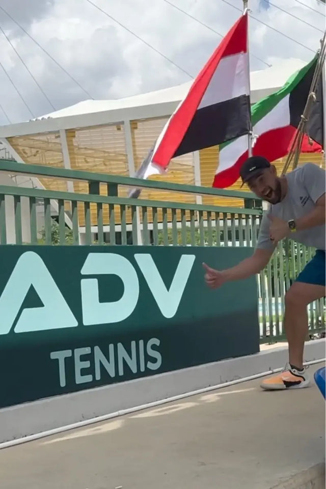 Grinning player gives a thumbs up beside the ADV Tennis sign under national flags.