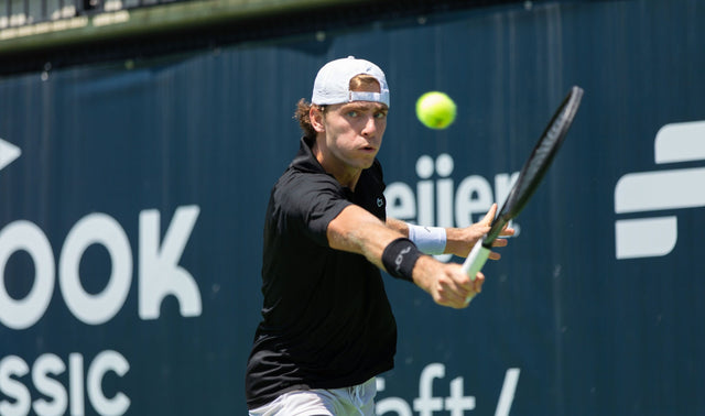 Tennis player in a backwards white cap tracking the ball for a two-handed backhand.