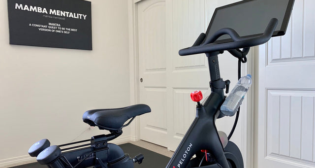 Peloton spin bike in a home gym beside a Mamba Mentality motivational wall sign.