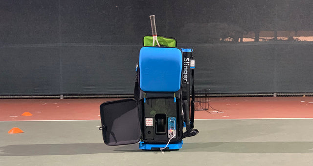 Blue Slinger Bag portable tennis ball machine standing upright on a court at night.