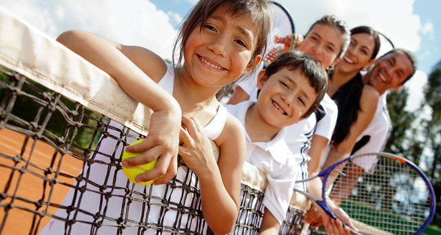 Family with two children leaning on a tennis net holding rackets on a clay court.