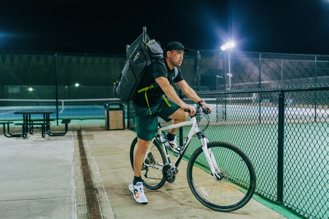 Cyclist wears an ADV tennis backpack beside a floodlit court at night.