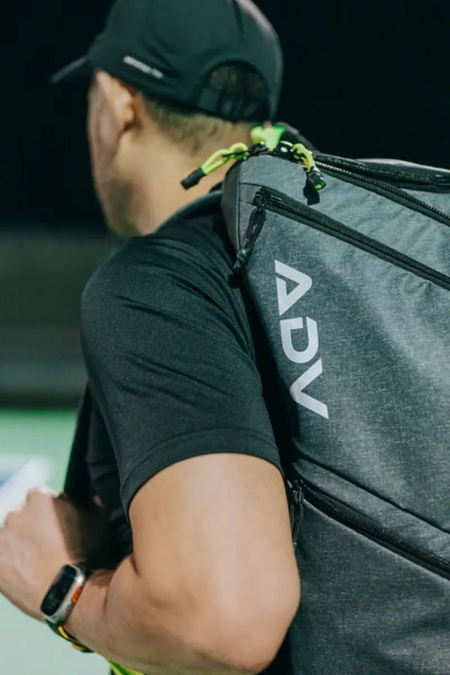 Player carrying a grey ADV tennis backpack with multiple zippered compartments on court.