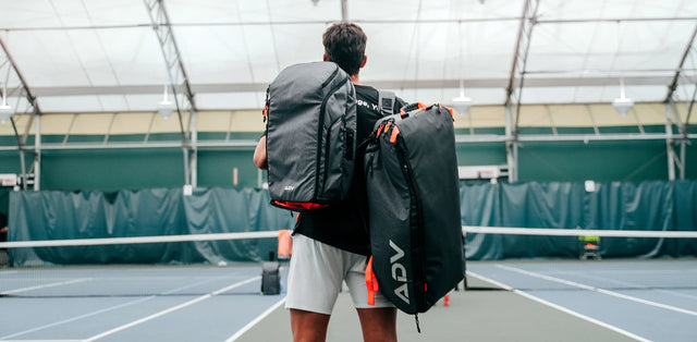 Player carrying two ADV tennis bags with orange trim on an indoor court.