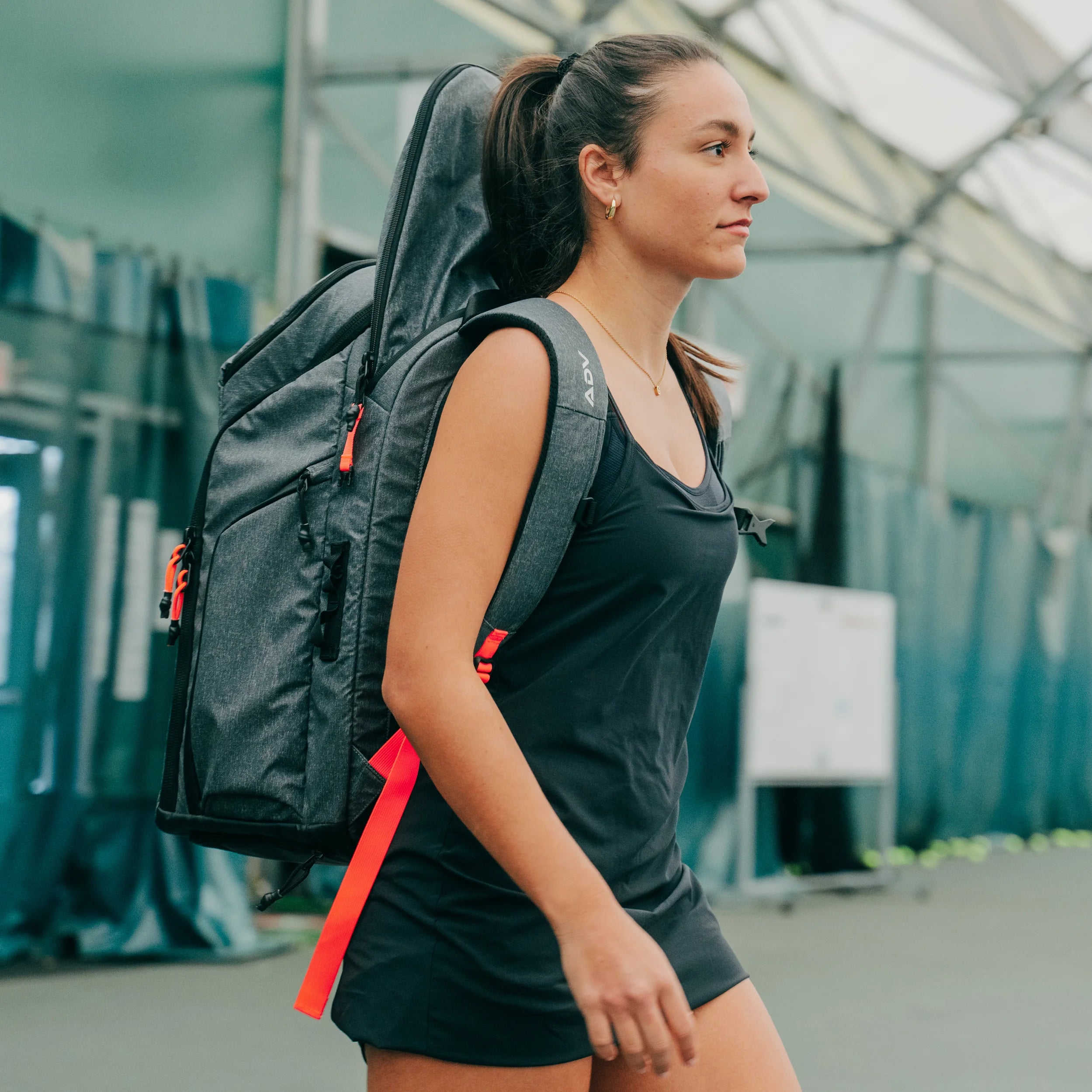 A woman carrying a gray 3 racket tennis bag with red accents on her back, walking on an indoor tennis court.