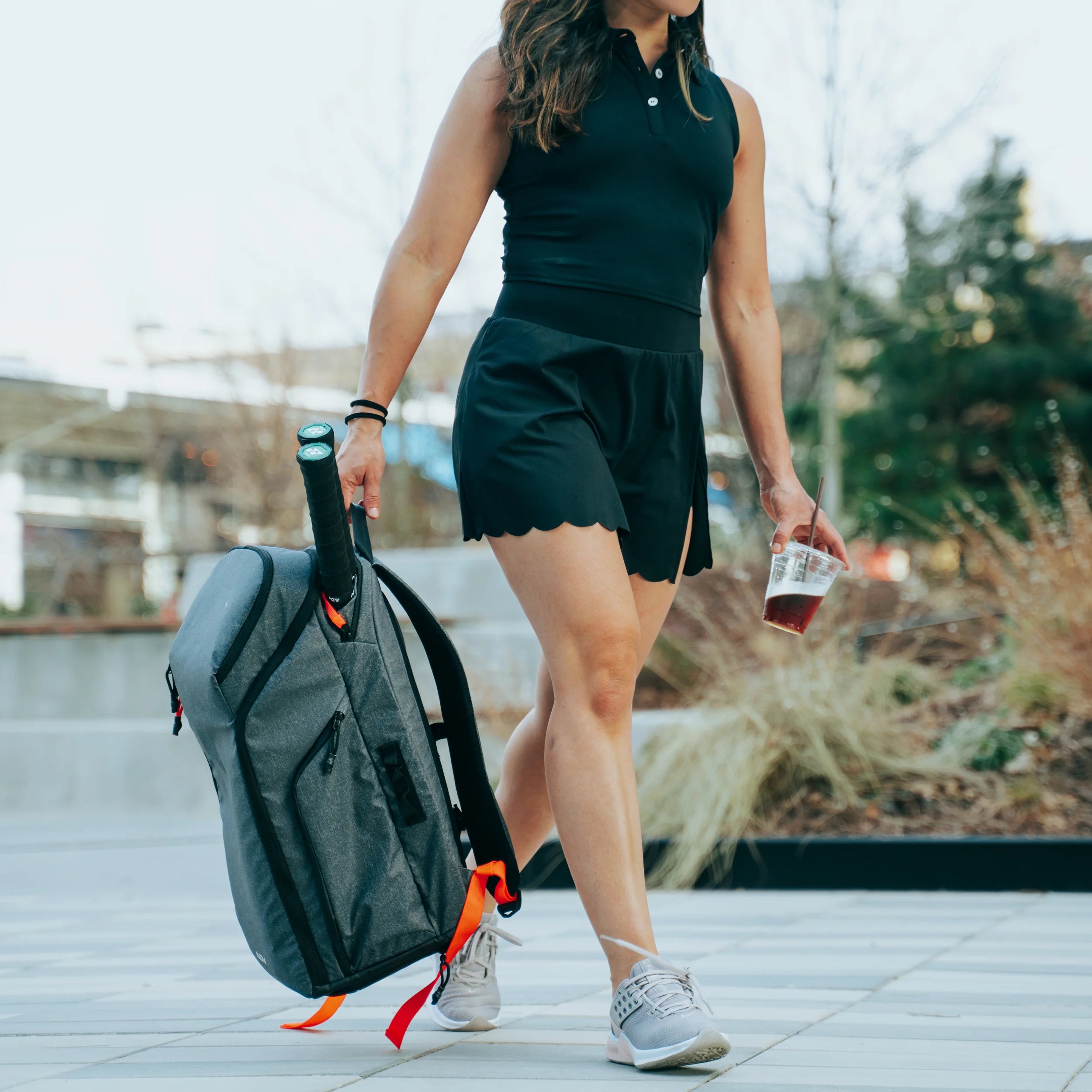 Gray tennis racket bag with red accents, carried by a woman walking outdoors while holding a drink.