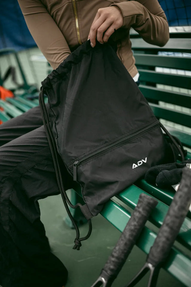 Person holding a black ADV Dash drawstring bag with zip pocket on a green bench.