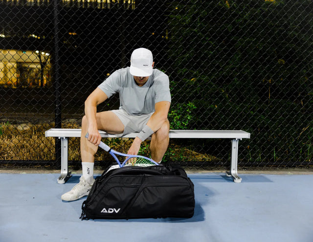 Player on a court bench packing a racquet into the black ADV Pro V3 tennis bag.