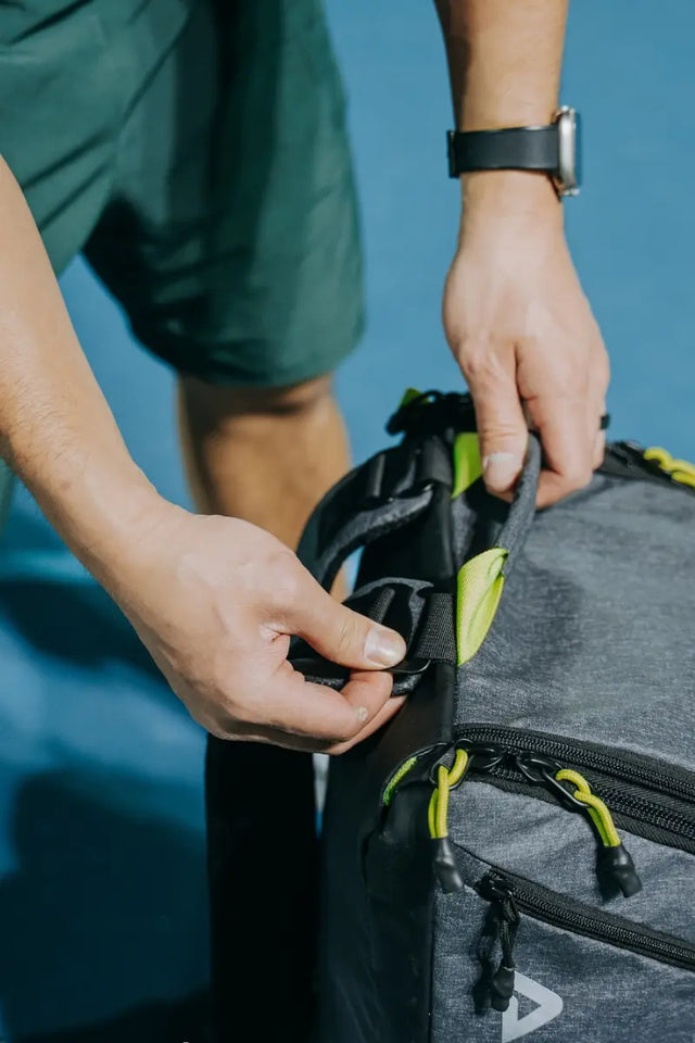 Hands adjusting straps on a grey tennis bag with green zippers against a blue wall.