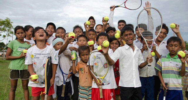 Smiling children in Cambodia holding up tennis rackets and balls at an outdoor court.
