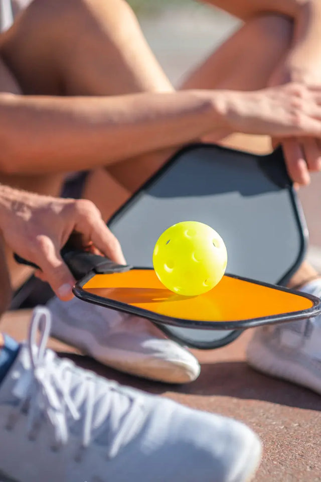 Pickleball paddle with an orange surface balancing a bright yellow pickleball, held by a player sitting on the court