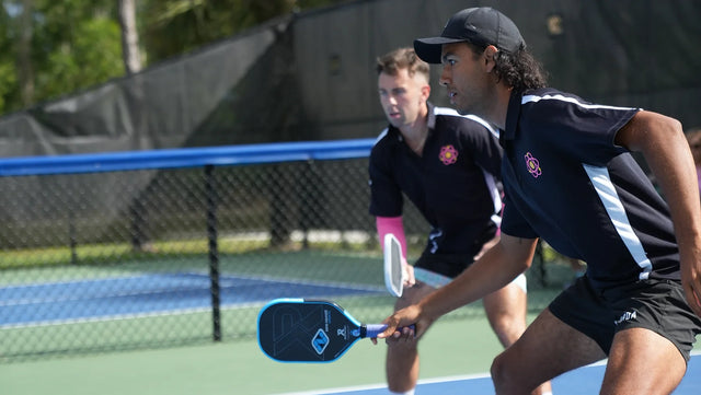 Pickleball players in black uniforms focused on a doubles match, with one holding a blue paddle mid-game.