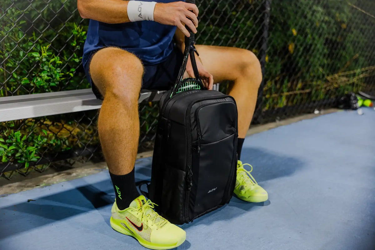 Player carrying two ADV tennis bags with orange trim on an indoor court.
