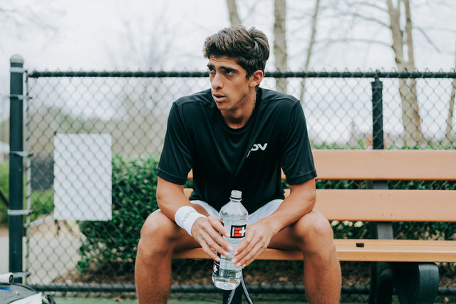 Tennis player in black ADV shirt resting on courtside bench holding a water bottle.