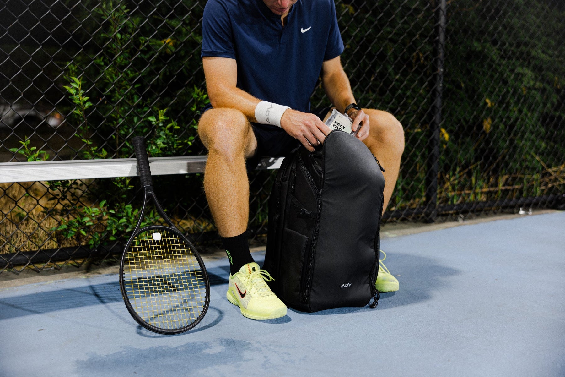 Player in ADV black shirt leaping to serve on a blue outdoor tennis court.