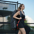 Tennis backpack carried by a woman in tennis attire walking beside an outdoor court.