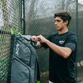 Pro tennis bag in gray with green accents, shown being unzipped by a player next to a court fence.
