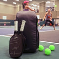 ADV Pickleball Backpack V2 in gray leaning against paddle cases on an indoor pickleball court, with balls nearby