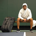 Tennis racket backpack resting upright on indoor court beside athlete seated on bench in white hoodie and black sneakers.