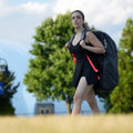 Tournament tennis backpack carried by a female player walking outdoors in tennis apparel with trees and sky in the background.