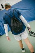 Tennis player carrying a black ADV drawstring bag over his shoulder while walking on court with a racket.