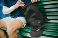 ADV black drawstring tennis bag and cap resting on a green bench beside a seated player.
