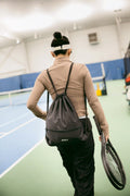 Female tennis player walking onto an indoor court with a black ADV drawstring bag and racket in hand.