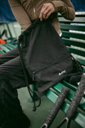 Close-up of a black ADV drawstring tennis bag with zippered pocket held courtside on green bleachers.