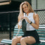 Smiling female tennis player sitting on bleachers wrapping white grip tape around her tennis racket handle