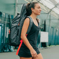 A woman carrying a gray 3 racket tennis bag with red accents on her back, walking on an indoor tennis court.