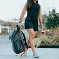 Gray tennis racket bag with red accents, carried by a woman walking outdoors while holding a drink.