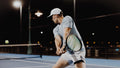 Tennis player in ADV cap and wristband hitting a backhand on a floodlit court at night.