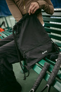 Person holding a black ADV Dash drawstring bag with zip pocket on a green bench.