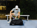 Player on a court bench packing a racquet into the black ADV Pro V3 tennis bag.