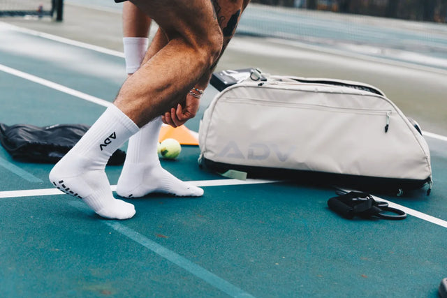 White ADV Pro Socks worn on a tennis court beside a duffel bag and ball.